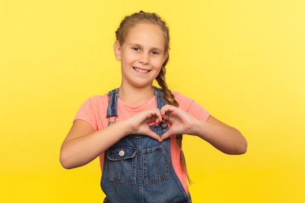 Foto porträt eines entzückenden kleinen mädchens mit zopf in denim-overalls, das herzform mit fingern zeigt, die unschuldige kindheitsliebesfreundschaft ausdrücken, studioaufnahme isoliert auf gelbem hintergrund