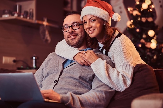 Porträt eines entzückenden Paares mit Laptop - bezaubernde Frau in Weihnachtsmütze, die ihren Mann umarmt und in die Kamera schaut. Weibchen, das Heiligabend feiert.