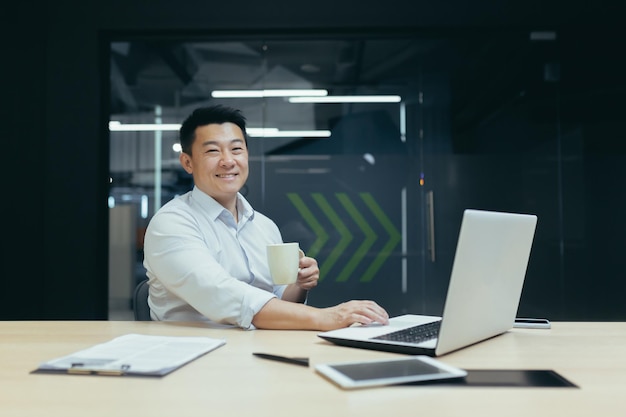 Porträt eines erfolgreichen asiatischen Chefs mit einer Tasse heißem Getränk, der lächelnd und in einem modernen Büro arbeitet