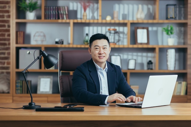 Porträt eines erfolgreichen asiatischen Geschäftsmannes, der im Büro arbeitet und am Tisch sitzt, in die Kamera blickt und einen glücklichen Banker mit Laptop lächelt