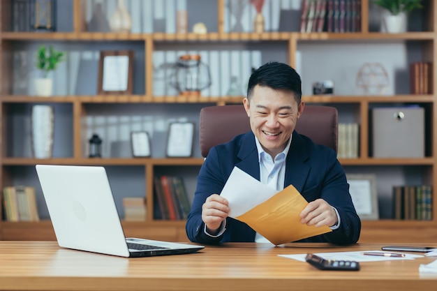 Porträt eines erfolgreichen asiatischen Geschäftsmannes erhielt von der Bank ein Genehmigungsschreiben, um die Kreditlinie zu verlängern. Ein glücklicher Mann, der am Tisch arbeitete und einen Umschlag hielt