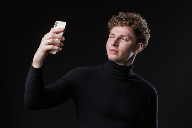 Porträt eines erfolgreichen jungen attraktiven, lockigen Geschäftsmannes, der vor einer schwarzen Wand steht und ein Selfie macht