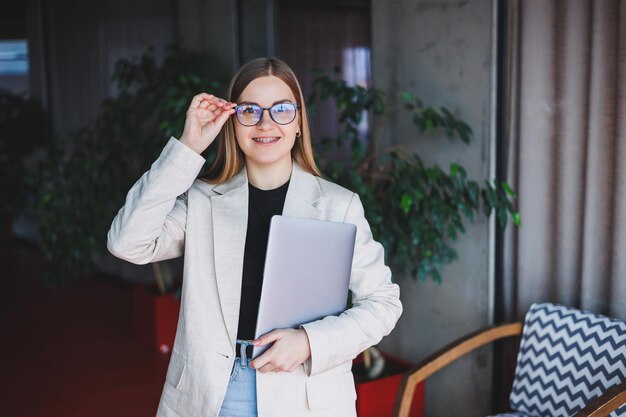 Porträt eines erfolgreichen Managers im Büro, der während des Arbeitstages hereinlächelt glückliche Frau mit Laptop, die Zeit im modernen Büro genießt Blondhaired Frau in einer beigen Jacke und Brille