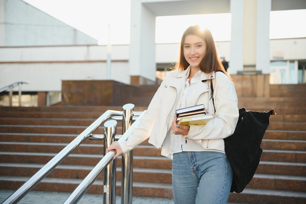 Porträt eines erfolgreichen Schülers, der glückliche Teenager-Mädchen posiert, die Bücher halten und lächelnd in die Kamera schauen, die in der Nähe des modernen Universitätsgebäudes außerhalb der Studentenschaft steht. Moderne Bildung