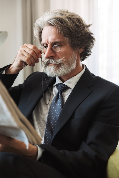 Porträt eines ernsten grauhaarigen leitenden Geschäftsmannes mit Brille zu Hause in formeller Kleidung, die Zeitung liest.