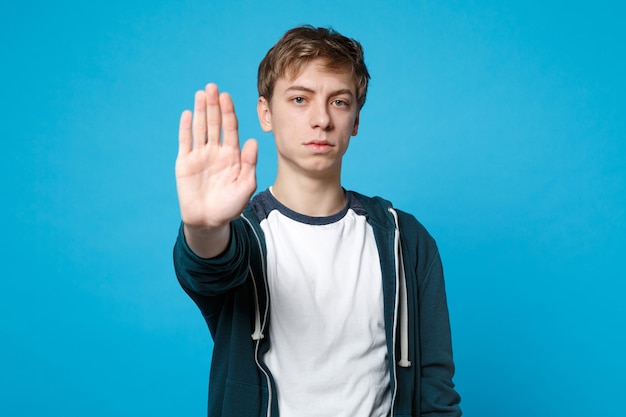 Porträt eines ernsten jungen Mannes in Freizeitkleidung, der eine Stoppgeste mit der Handfläche zeigt, die auf Blau isoliert ist. Menschen aufrichtige Emotionen, Lifestyle-Konzept.