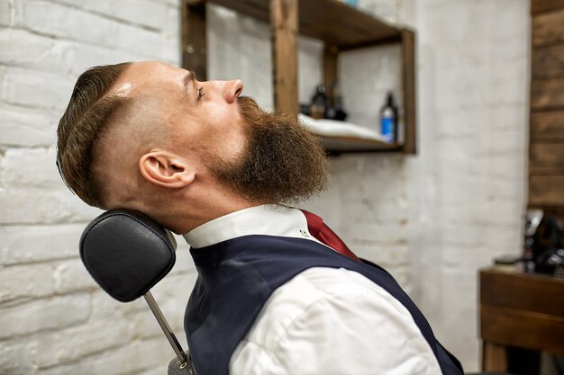 Porträt eines ernsten Mannes in einem Friseurladen. Ein Mann sitzt auf dem Friseurstuhl. Kopieren Sie Platz auf Backsteinmauerhintergrund.