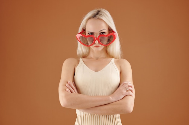 Porträt eines ernsten schönen kaukasischen Mädchens in herzförmiger Brille und Kleid, das mit verschränkten Armen vor braunem Hintergrund steht