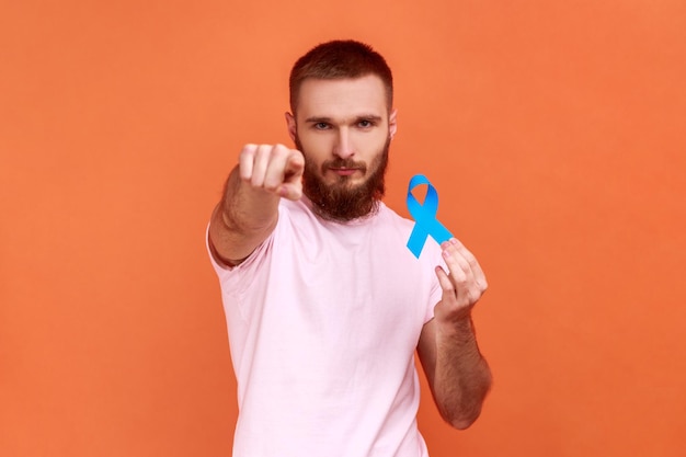 Porträt eines ernsthaften bärtigen Mannes, der mit dem Finger auf die Kamera zeigt und ein blaues Bewusstseinskrankheitssymbol hält, das die Kameraunterstützung mit rosafarbenem T-Shirt anschaut Innenstudioaufnahme isoliert auf orangefarbenem Hintergrund