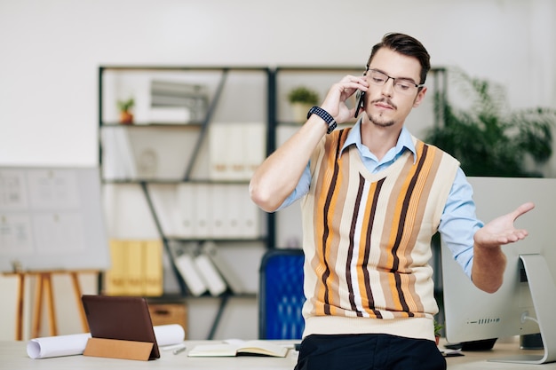 Porträt eines ernsthaften jungen Unternehmers mit Brille, der beim Telefonieren mit einem Kollegen gestikuliert