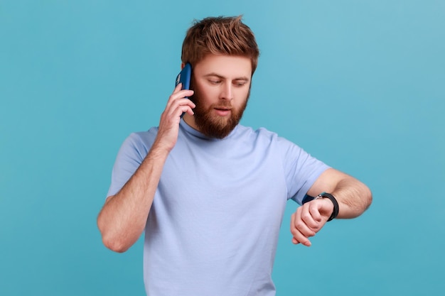 Porträt eines ernsthaften, konzentrierten bärtigen Mannes, der am Telefon spricht und die Zeit auf seinen Armbanduhren sieht, die sagen, dass er zu spät zum Treffen kommt Studioaufnahme im Innenbereich isoliert auf blauem Hintergrund