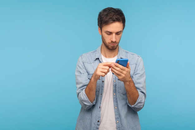 Porträt eines ernsthaften Mannes im Arbeiter-Jeanshemd, der eine SMS-Nachricht in sozialen Medien auf dem Handy unter Verwendung von Mobilfunknetzdiensten sendet, die online im Innenbereich chatten, Studioaufnahme isoliert auf blauem Hintergrund, Kopienraum