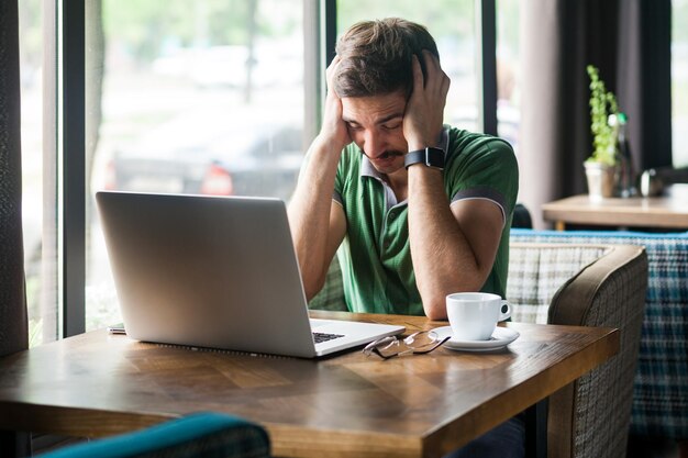 Porträt eines erschöpften, müden Freiberuflers mit grünem T-Shirt, der an Migräne oder Bluthochdruck leidet, während er online am Laptop arbeitet Innenaufnahme in der Nähe eines großen Fenstercafés Hintergrund