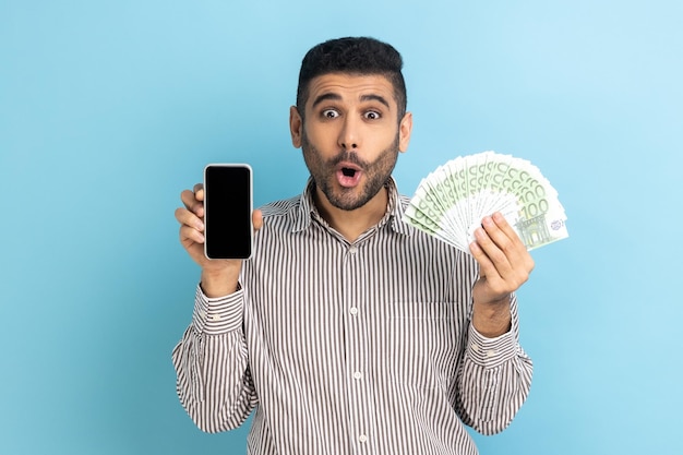 Porträt eines erstaunten Geschäftsmannes, der einen großen Fan von Euro-Banknoten und ein Mobiltelefon mit leerem Display für Werbung mit gestreiftem Hemd hält. Studioaufnahme im Innenbereich isoliert auf blauem Hintergrund