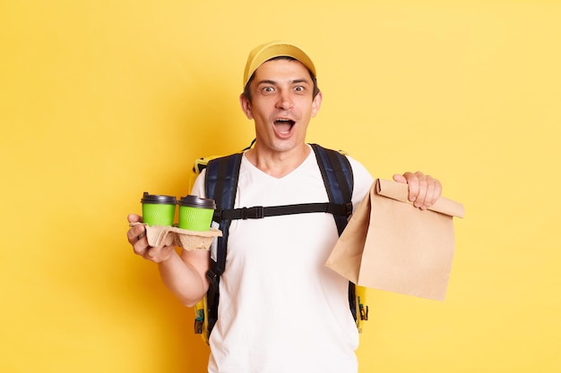 Porträt eines erstaunten Lieferboten mit weißem T-Shirt und Mütze, der ein Papierpaket hält und Kaffee in den Händen mitnimmt. Schneller Lieferkurier, der isoliert auf gelbem Hintergrund posiert