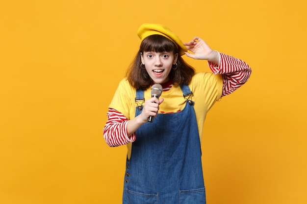 Porträt eines erstaunten Teenagers in französischer Baskenmütze, Denim-Sommerkleid singt Lied im Mikrofon isoliert auf gelbem Wandhintergrund im Studio. Menschen aufrichtige Emotionen, Lifestyle-Konzept. Mock-up-Kopienbereich.