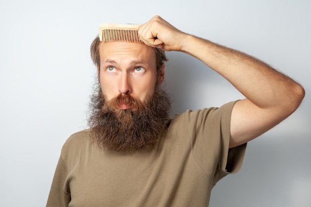 Porträt eines erwachsenen glatzköpfigen Mannes mit Haarbürste, der an Alopezie leidet und mit verwirrtem Gesichtsausdruck nach oben schaut bärtiger Mann mit T-Shirt Studioaufnahme im Innenbereich isoliert auf grauem Hintergrund