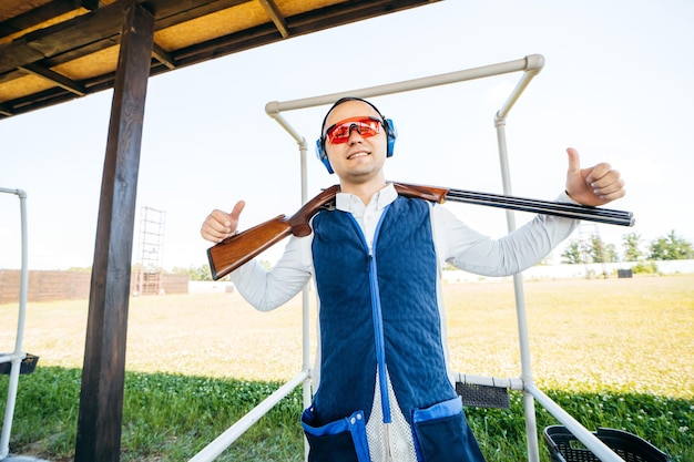 Porträt eines erwachsenen lächelnden Mannes mit Sonnenbrille, schützenden Kopfhörern und einer Gewehrweste mit Schrotflinte