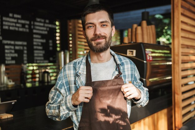 Porträt eines europäischen Barista-Typs, der eine Schürze trägt und lächelt, während er im Straßencafé oder Kaffeehaus im Freien arbeitet?