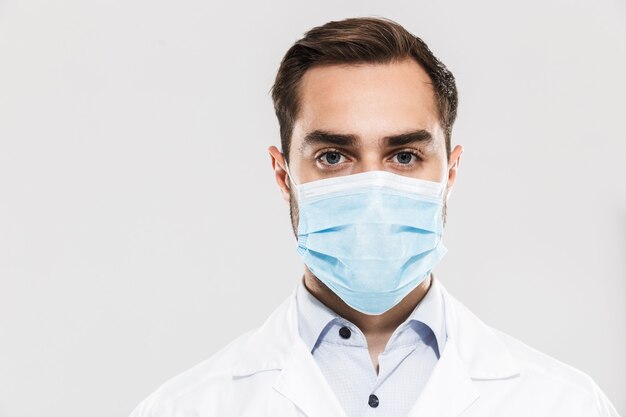 Porträt eines europäischen jungen Arztes mit steriler Maske, der im Kliniklabor arbeitet, isoliert über weißer Wand