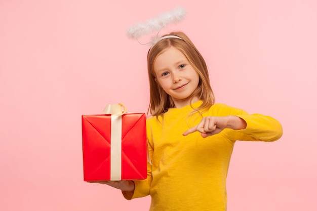 Porträt eines faszinierenden kleinen rothaarigen Mädchens mit Sommersprossen und engelhaftem Heiligenschein, das auf eine Geschenkbox zeigt, die zum Urlaub gratuliert und Geburtstagsgeschenk gibt, Studioaufnahme isoliert auf rosa Hintergrund