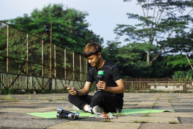 Porträt eines fitten jungen Mannes, der draußen auf einer Yogamatte sitzt und das Telefon und die Flasche hält