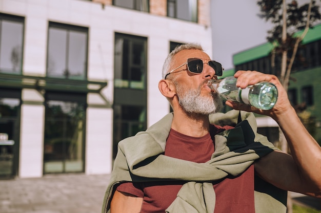 Porträt eines fitten reifen Mannes mit Sonnenbrille, der draußen Wasser aus der Glasflasche trinkt