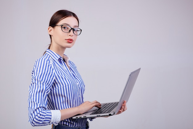 Porträt eines Frauenadministrators in einem gestreiften weiß-blauen Hemd mit Gläsern und einem Laptop auf Grau. Mitarbeiterin des Jahres, Business Lady.