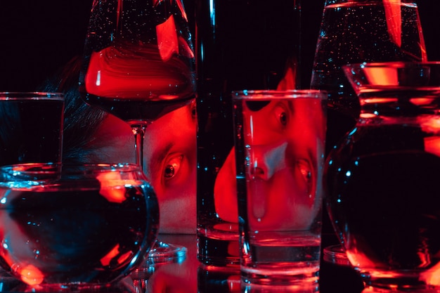 Porträt eines fremden Mannes, der durch Glasgläser des Wassers mit Reflexionen und Verzerrungen mit rotem blauem Neonlicht schaut