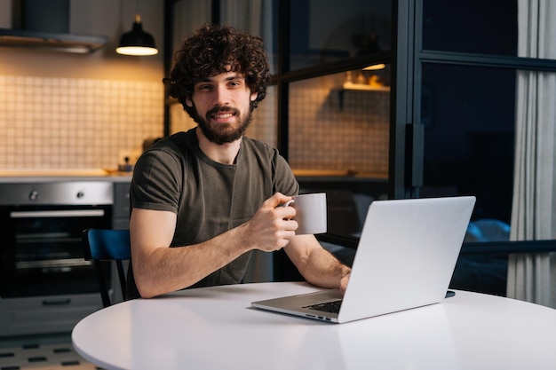 Porträt eines freundlichen bärtigen jungen Freiberuflers, der in der Hand eine Tasse mit Morgenkaffee hält, der am Tisch mit einem Laptop sitzt, der in die Kamera blickt