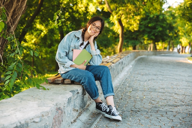 Porträt eines fröhlich lächelnden süßen jungen Studentenmädchens mit Brille, das draußen auf einer Bank im Naturpark sitzt und Bücher hält.