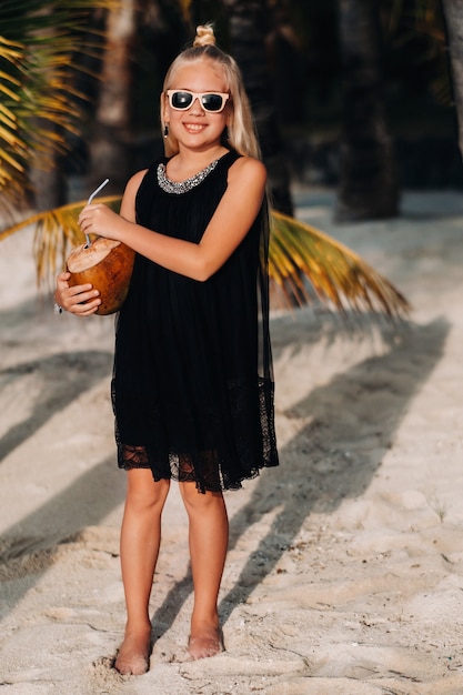 Porträt eines fröhlichen 9-jährigen Mädchens mit einem Kokosnusscocktail auf dem Hintergrund von Palmen an einem exotischen Strand. Kleines Mädchen mit einer Kokosnuss am Strand der Insel Mauritius.