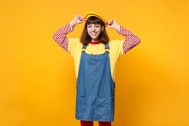 Porträt eines fröhlichen, charmanten Mädchen-Teenagers im Denim-Sommerkleid, das die Hände auf französischer Baskenmütze hält, isoliert auf gelbem Wandhintergrund im Studio. Menschen aufrichtige Emotionen Lifestyle-Konzept. Mock-up-Kopienbereich.