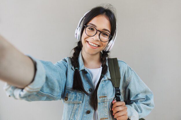 Porträt eines fröhlichen, glücklichen, schönen Mädchens in Jeansjacke, das eine Brille trägt, die über grauer Wand isoliert ist und Musik mit Kopfhörern hört, macht ein Selfie mit der Kamera.