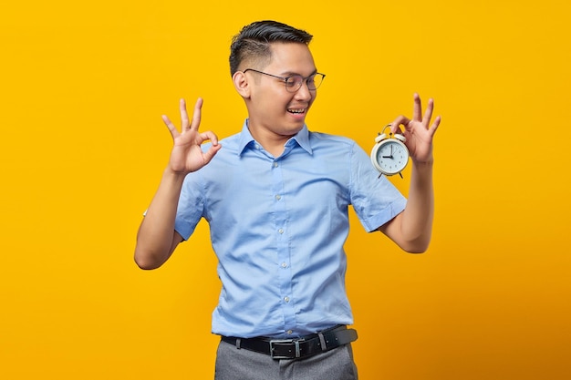 Porträt eines fröhlichen, gutaussehenden asiatischen jungen Mannes mit Brille, der einen Wecker zeigt und eine Ok-Geste mit dem Finger zeigt, der auf gelbem Hintergrund isoliert ist Zeitmanagement-Lifestyle-Konzept