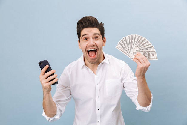 Porträt eines fröhlichen gutaussehenden jungen Mannes, der isoliert auf Blau steht und Geldbanknoten zeigt, die Handy halten