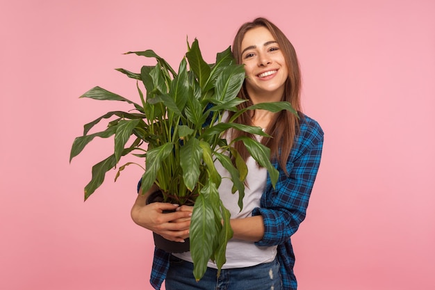 Porträt eines fröhlichen Hausfrauenmädchens in kariertem Hemd, das mit erfreutem Lächeln steht und einen Blumentopf hält, der grüne große Pflanzen bewundert, liebt die Gartenarbeit im Innenbereich, Studioaufnahme isoliert auf rosa Hintergrund