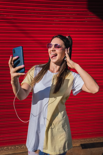 Porträt eines fröhlichen hübschen Mädchens, das Musik mit Kopfhörern hört, während es steht und ein Selfie macht.