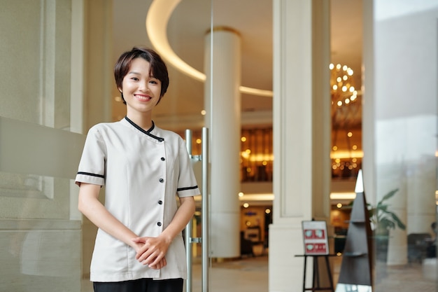 Porträt eines fröhlichen jungen asiatischen Hotelmanagers, der an den Eingangstüren steht und lächelt