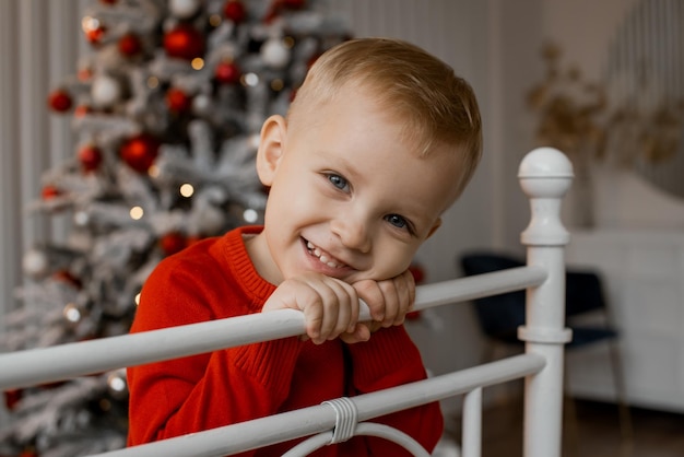 Porträt eines fröhlichen Jungen in rotem Strickpullover, der auf dem Bett sitzt und neben dem Weihnachtsbaum zu Hause in die Kamera lächelt Familien und Kinder feiern Winterferien