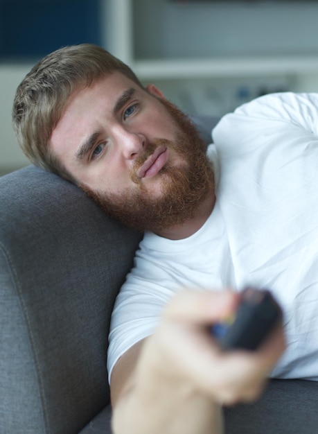 Porträt eines fröhlichen jungen Mannes in legerer Kleidung, der auf dem Sofa im Wohnzimmer sitzt und fernsieht