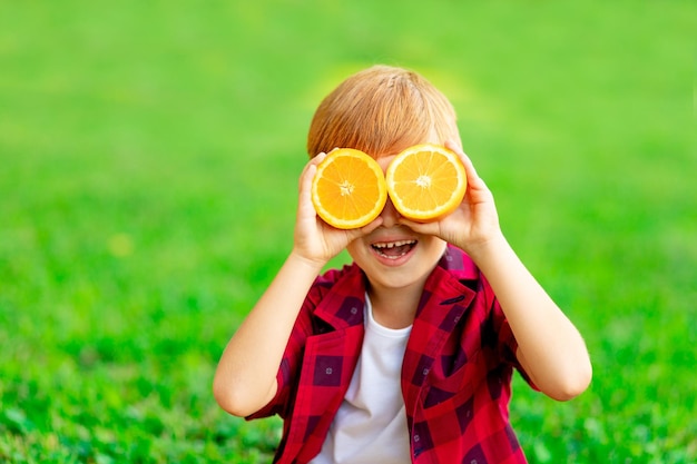 Porträt eines fröhlichen kleinen rothaarigen Jungen in einem roten Hemd auf einem grünen Rasen im Sommer, der fröhlich lächelt oder lacht, während er seine Augen mit einer Orange schließt