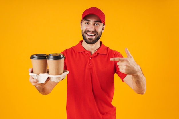 Porträt eines fröhlichen Lieferboten in roter Uniform, der Kaffeetassen zum Mitnehmen isoliert über Gelb lächelt und hält