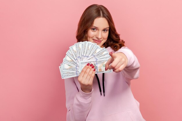 Porträt eines fröhlichen, lockigen Teenager-Mädchens, das einen Haufen Dollar-Banknoten hält und mit dem Finger auf die Kamera zeigt, hey du Geste, Gewinner. Studioaufnahme im Innenbereich isoliert auf rosa Hintergrund