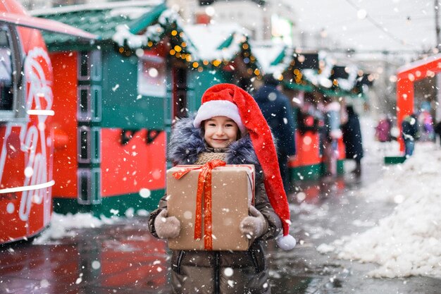 Porträt eines fröhlichen Mädchens in Weihnachtsmütze mit Geschenkbox für Weihnachten auf der Stadtstraße im Winter mit Schnee auf festlichem Markt mit Dekorationen und Lichterketten. Warme Kleidung, Strickschal und Pelz. Neujahr