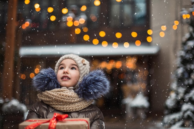 Porträt eines fröhlichen Mädchens mit einer Geschenkbox zu Weihnachten auf einer Stadtstraße im Winter mit Schnee auf einem festlichen Markt mit Dekorationen und Lichterketten Warme Kleidung Strickmütze Schal und Fell Neues Jahr