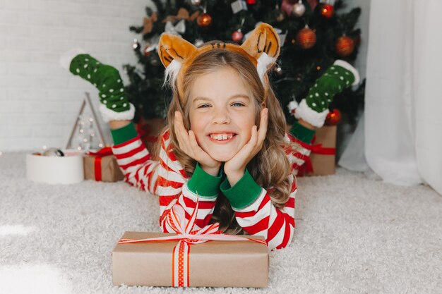 Porträt eines fröhlichen Mädchens von 9 Jahren, das auf dem Boden in der Nähe von Kisten mit Geschenken und einem Weihnachtsbaum zu Hause liegt. ein Mädchen in einer Felge mit Tigerohren.