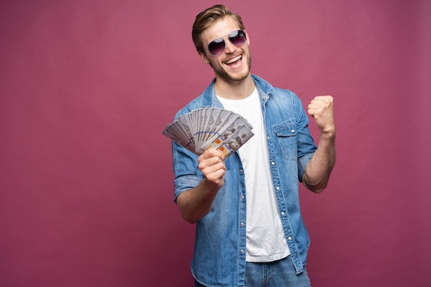 Porträt eines fröhlichen Mannes in Jeanshemd, der einen Haufen Geldbanknoten hält, während er steht und auf rosafarbenem Hintergrund isoliert feiert