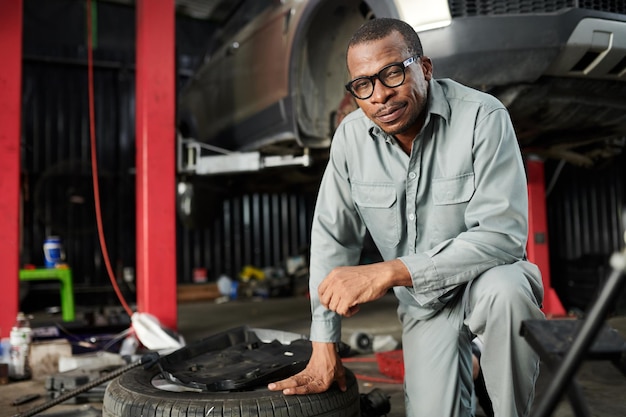 Foto porträt eines fröhlichen mechanikers in grauem overall, der sich auf einen reifen stützt und in die kamera lächelt