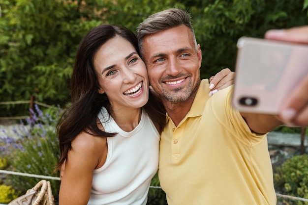 Porträt eines fröhlichen Paares mittleren Alters, das ein Selfie-Foto auf dem Handy macht und sich beim Gehen im Sommerpark umarmt